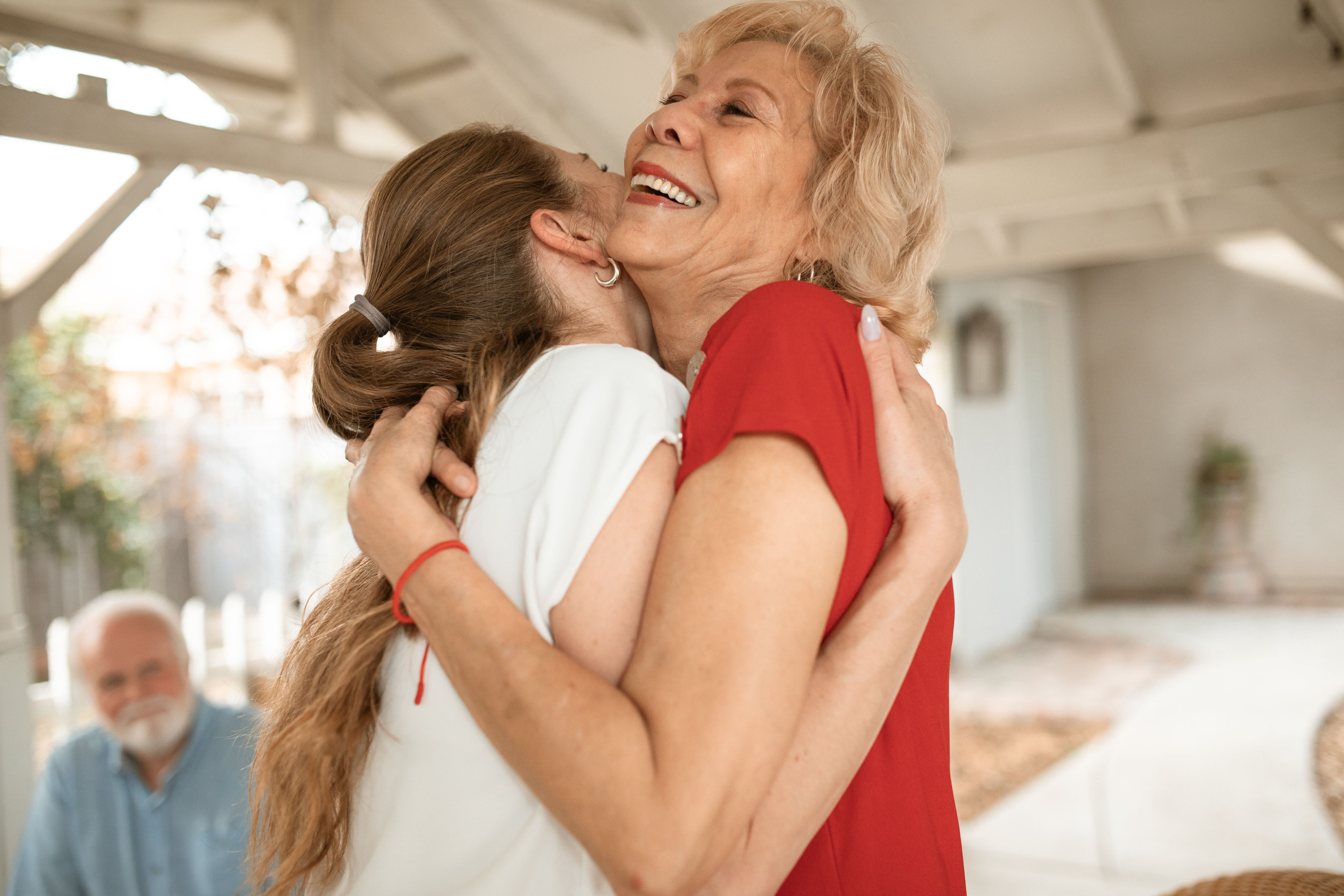 A daughter and her mother embracing at home, an older man smiling behind them.